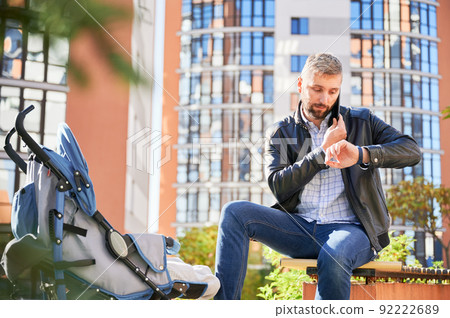 Father making business call, looking on watch while spending time with son outdoors. Front view of bearded man holding mobile phone, calling, while baby sleeping in pram in park. Concept of working. 92222689