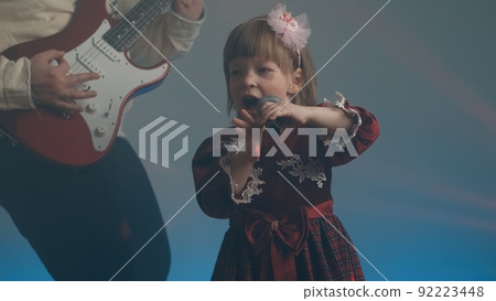 Little girl on stage in vintage dress, she sings into microphone and dances, her father plays an electric guitar. Color music is shining and smoke is billowing. Family time, creativity and hobbies Little girl on stage in vintage dress, she sings into microphone and dances, her father plays an electric guitar. Color music is shining and smoke is billowing. Family time, creativity and hobbies 92223448