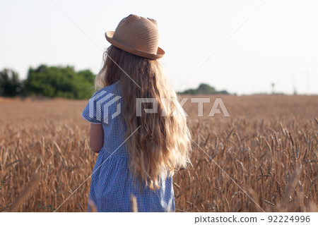 Adorable little girl in a straw hat, blue plaid summer dress in wheat field countryside.Child with long blonde wavy hair Adorable little girl in a straw hat, blue plaid summer dress in wheat field countryside.Child with long blonde wavy hair 92224996