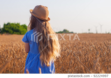 Adorable little girl in a straw hat, blue plaid summer dress in wheat field countryside.Child with long blonde wavy hair Adorable little girl in a straw hat, blue plaid summer dress in wheat field countryside.Child with long blonde wavy hair 92224997
