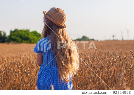 Adorable little girl in a straw hat, blue plaid summer dress in wheat field countryside.Child with long blonde wavy hair Adorable little girl in a straw hat, blue plaid summer dress in wheat field countryside.Child with long blonde wavy hair 92224998