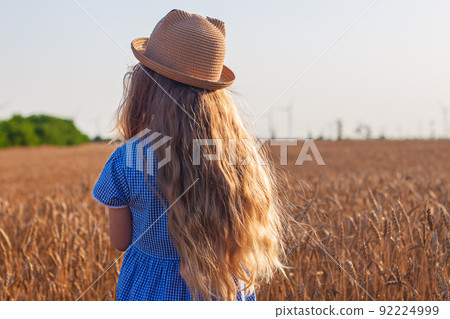 Adorable little girl in a straw hat, blue plaid summer dress in wheat field countryside.Child with long blonde wavy hair Adorable little girl in a straw hat, blue plaid summer dress in wheat field countryside.Child with long blonde wavy hair 92224999