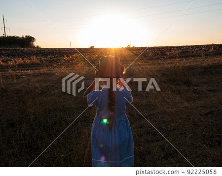 Adorable little girl in a straw hat, blue plaid summer dress in grass field countryside. Child with long blonde braid. 92225058