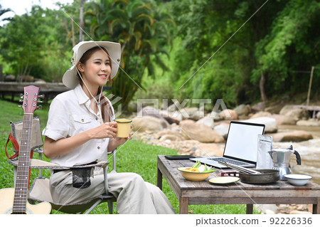 Happy female traveler drinking coffee, resting in folding chair near the river bank. Adventure, travel and camping concept 92226336