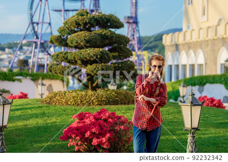 A woman in a youth shirt and jeans poses standing by a flower bed against the backdrop of an amusement park and a cityscape in the sunset A woman in a youth shirt and jeans poses standing by a flower bed against the backdrop of an amusement park and a cityscape in the sunset 92232342