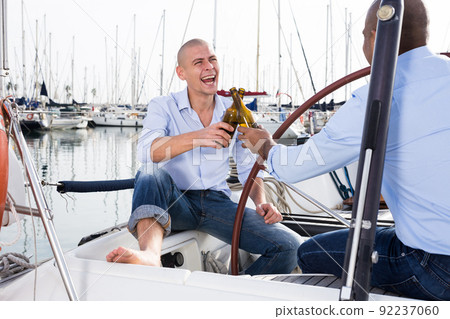 Two men in blue shirts and jeans with bare legs drinking beer and having fun on a yacht in seaport 92237060