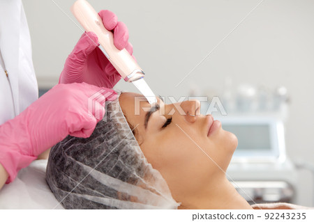 Cosmetologist, beautician in pink gloves making facial treatment with ultrasonic spatula to woman, face skin scrubber treatment with ultrasonic spatula, facial cleansing procedure in beauty salon 92243235