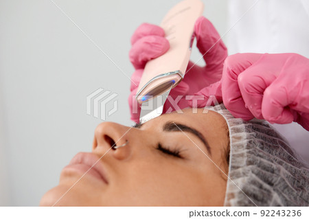 Cosmetologist, beautician in pink gloves making facial treatment with ultrasonic spatula to woman, face skin scrubber treatment with ultrasonic spatula, facial cleansing procedure in beauty salon 92243236
