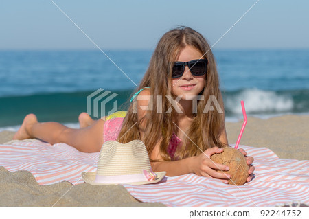 A teenage girl with a coconut lies on a beach towel on the seashore. 92244752