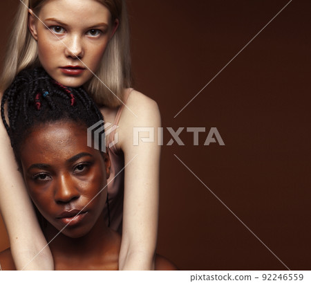two pretty girls african and caucasian blond posing cheerful together on brown background, ethnicity diverse lifestyle people concept close up 92246559
