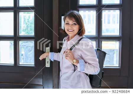 Middle aged woman looking at camera while standing near front door to house Middle aged woman looking at camera while standing near front door to house 92248684