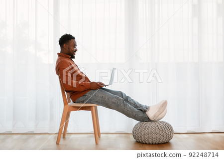 Cheerful African American Guy Using Laptop Sitting In Chair Indoors Cheerful African American Guy Using Laptop Sitting In Chair Indoors 92248714