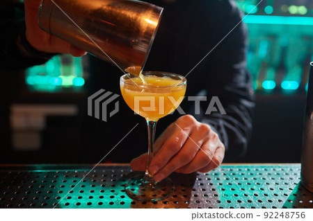 Barman in shirt and apron making an alcoholic drink with ice in a cocktail glass. Barman in shirt and apron making an alcoholic drink with ice in a cocktail glass. 92248756