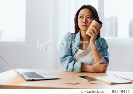 Upset frustrated pensive young Asian cute female businesswoman reclines on coffee cup looks aside in light office interior. Freelancer work at home. Lady corporation leader concept. Copy space Offer Upset frustrated pensive young Asian cute female businesswoman reclines on coffee cup looks aside in light office interior. Freelancer work at home. Lady corporation leader concept. Copy space Offer 92249455