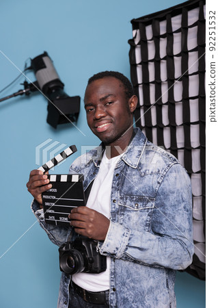Smiling photographer with clapper on blue background while having softboxes and spotlights. Young confident producer with clapperboard standing in studio having professional production equipment. Smiling photographer with clapper on blue background while having softboxes and spotlights. Young confident producer with clapperboard standing in studio having professional production equipment. 92251532