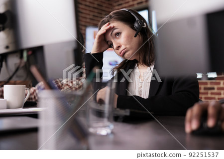 Young woman feeling tired at customer care support job, working on telemarketing call center at helpdesk. Female receptionist with helpline service helping clients on remote communication. 92251537