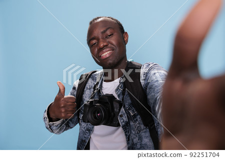 Photography enthusiast giving approve hand gesture while taking picture of himself. Confident photographer showing okay finger symbol while taking selfie and having DSLR camera on blue background. Photography enthusiast giving approve hand gesture while taking picture of himself. Confident photographer showing okay finger symbol while taking selfie and having DSLR camera on blue background. 92251704