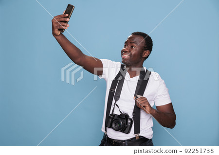 Cheerful photographer taking selfie photo with smartphone while smiling heartily on blue background. Confident and handsome photography enthusiast taking picture of himself while having DSLR camera. Cheerful photographer taking selfie photo with smartphone while smiling heartily on blue background. Confident and handsome photography enthusiast taking picture of himself while having DSLR camera. 92251738