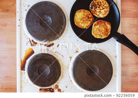 dirty kitchen stove with stains of burnt food and fat, frying pan with pancakes on top 92256059
