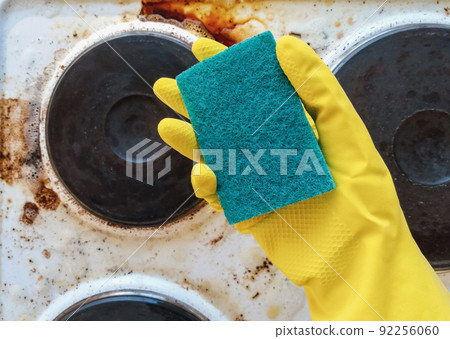 hand in yellow glove holding washup sponge against dirty electric stove hand in yellow glove holding washup sponge against dirty electric stove 92256060