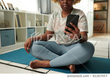 Close-up of African young woman sitting on exercise mat and watching sports training online using her mobile phone Close-up of African young woman sitting on exercise mat and watching sports training online using her mobile phone 92267857