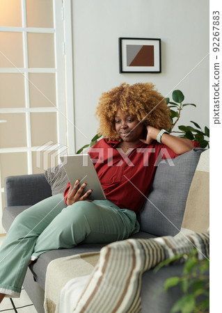 African girl resting on sofa in living room and having online communication on digital tablet African girl resting on sofa in living room and having online communication on digital tablet 92267883