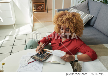 African young woman holding paper bill using calculator while sitting at coffee table with notepad and planning her budget African young woman holding paper bill using calculator while sitting at coffee table with notepad and planning her budget 92267959