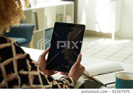 Close-up of African girl looking at screen of digital tablet and working online sitting at table with notepad in the room 92267970