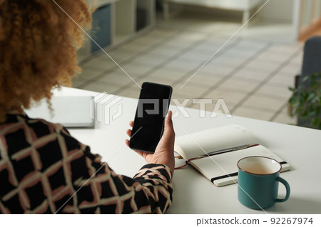 Close-up of African girl looking at screen of her mobile phone while sitting at table with notepad and cup of coffee Close-up of African girl looking at screen of her mobile phone while sitting at table with notepad and cup of coffee 92267974