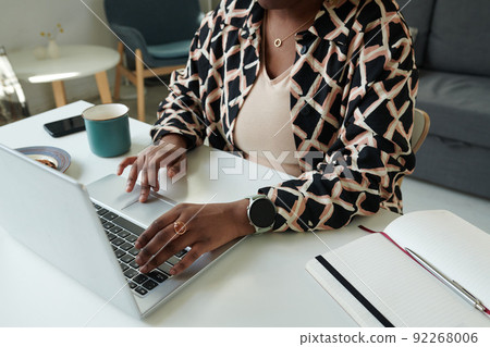 Close-up of African girl doing her online work typing on laptop while sitting at table with notepad and cup of coffee at home 92268006
