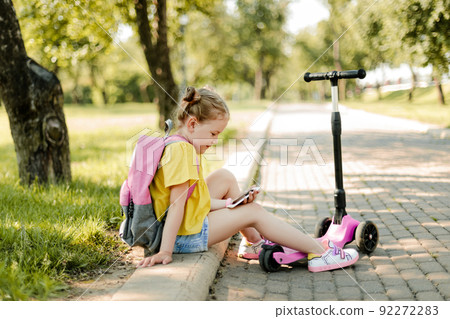 A smiling little schoolgirl girl is sitting on the curb with a phone in her hands during a break between lessons. Modern technology, back to school A smiling little schoolgirl girl is sitting on the curb with a phone in her hands during a break between lessons. Modern technology, back to school 92272283