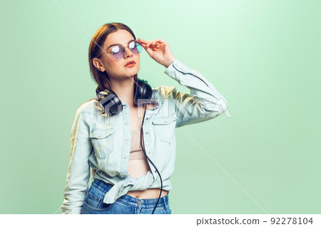 Portrait of young girl, student in casual clothes, sunglasses and headphones posing isolated over green studio background. 92278104