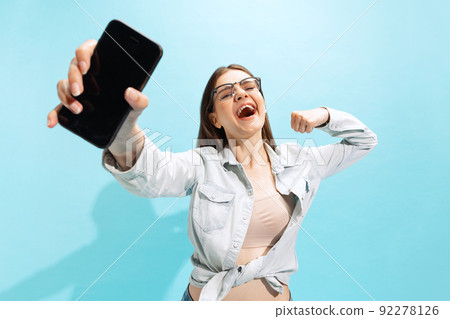 Portrait of happy young girl in casual clothes showing phone isolated over blue studio background. Winning lotey 92278126
