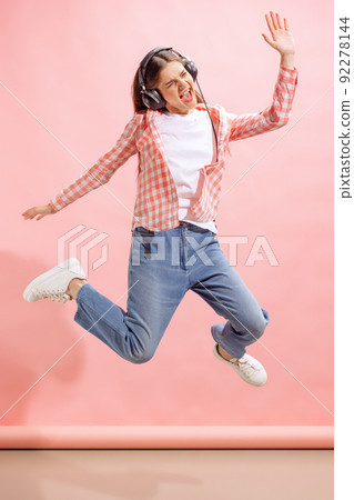 Portrait of emotive young girl in checkered shirt listening to music in headphones and jumping isolated over pink studio background Portrait of emotive young girl in checkered shirt listening to music in headphones and jumping isolated over pink studio background 92278144