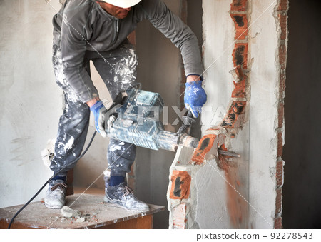 Close up of male builder in workwear drilling wall with hammer drill. Man worker using drill breaker while destroying wall in apartment under renovation. Demolition work and home renovation concept. 92278543