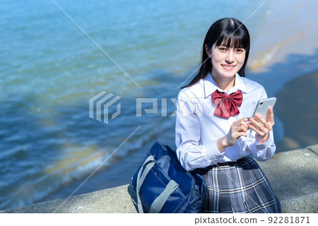 A high school girl app that sits on a beautiful beach breakwater and uses a smartphone SNS mobile phone After school at the beach 92281871