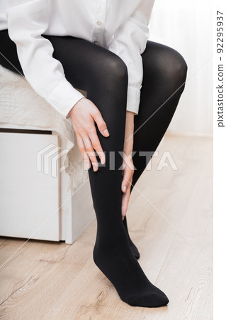 Girl putting on stockings at home. Black compression stockings on a woman in a white room. Black tights. Beautiful female legs. 92295937