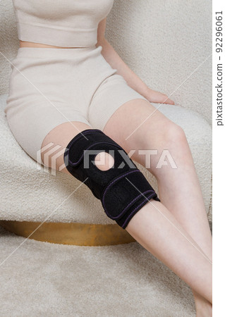 Black knee support brace on a woman leg. girl in an orthosis in the interior of the house. Orthopedic anatomic braces for knee fixation, injuries and pain. Knee Joint Bandage Sleeve. Elastic Sports. 92296061