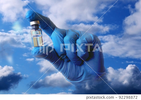 A hand in a blue medical glove sticks out of the clouds and holds out a vial of vaccine. Collage.. A hand in a blue medical glove sticks out of the clouds and holds out a vial of vaccine. Collage.. 92298872