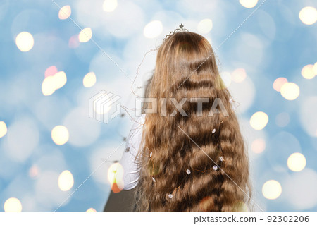 Rear view of a girl with long wavy flowing hair on a bright shimmering blue and yellow background. A crown and garlands of snowflakes for a child. Merry Christmas and Happy New Year. Copy space. 92302206