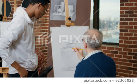 Young artist explaining drawing technique to elder person, using pencil to develop artistic skills on canvas and easel. Senior student learning to draw vase model in art class with teacher. Young artist explaining drawing technique to elder person, using pencil to develop artistic skills on canvas and easel. Senior student learning to draw vase model in art class with teacher. 92305028