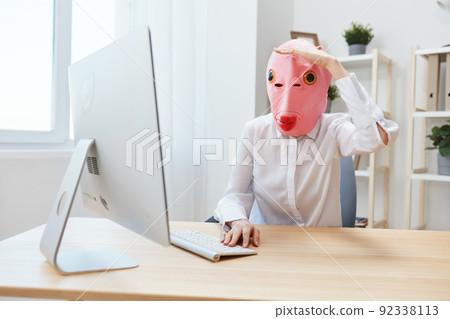 Funny crazy businesswoman worker freelancer in pink fish mask work on computer online pondering or making decision holds hand over eyes and looks into distance in light modern office. Copy space Funny crazy businesswoman worker freelancer in pink fish mask work on computer online pondering or making decision holds hand over eyes and looks into distance in light modern office. Copy space 92338113