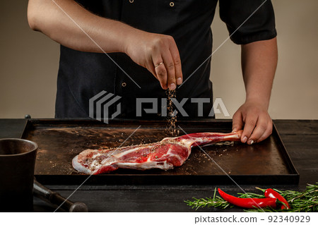 Male chef rubbing raw lamb shanks with salt and pepper on stone tray on wooden table. Chef cooking appetizing shank of lamb. Male chef rubbing raw lamb shanks with salt and pepper on stone tray on wooden table. Chef cooking appetizing shank of lamb. 92340929