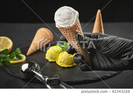 Hand in a dark glove is holding a waffle cone with a creamy ice cream on a background of a lemon sorbet decorated with mint. 92343288