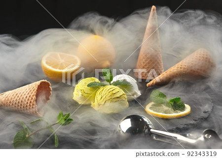 Tasty creamy and lemon ice cream decorated with mint served in smoke on a stone slate over a black background. Tasty creamy and lemon ice cream decorated with mint served in smoke on a stone slate over a black background. 92343319