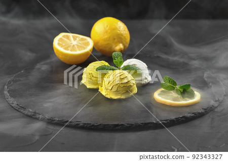 Tasty creamy and lemon ice cream decorated with mint served in smoke on a stone slate over a black background. Tasty creamy and lemon ice cream decorated with mint served in smoke on a stone slate over a black background. 92343327