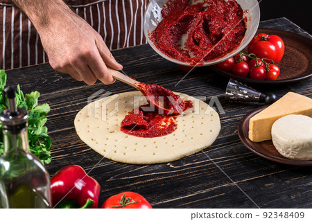 A male hand spreading tomato puree on a pizza base with spoon on an old wooden background A male hand spreading tomato puree on a pizza base with spoon on an old wooden background 92348409