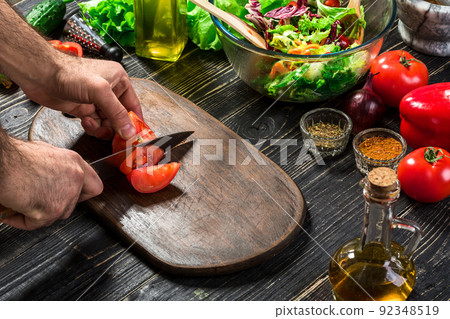 Man's hand cuts ripe red tomatoes for summer healthy vegetable salad on a wooden board, vertical Man's hand cuts ripe red tomatoes for summer healthy vegetable salad on a wooden board, vertical 92348519