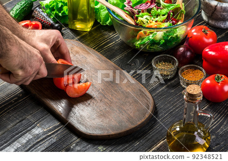 Man's hand cuts ripe red tomatoes for summer healthy vegetable salad on a wooden board, vertical Man's hand cuts ripe red tomatoes for summer healthy vegetable salad on a wooden board, vertical 92348521