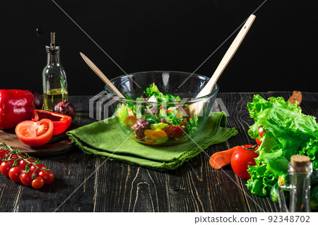 A glass bowl of fresh vegetable salad surrounded by the ingredients: green lettuce, cucumbers, sweet pepper and tomatoes. A glass bowl of fresh vegetable salad surrounded by the ingredients: green lettuce, cucumbers, sweet pepper and tomatoes. 92348702